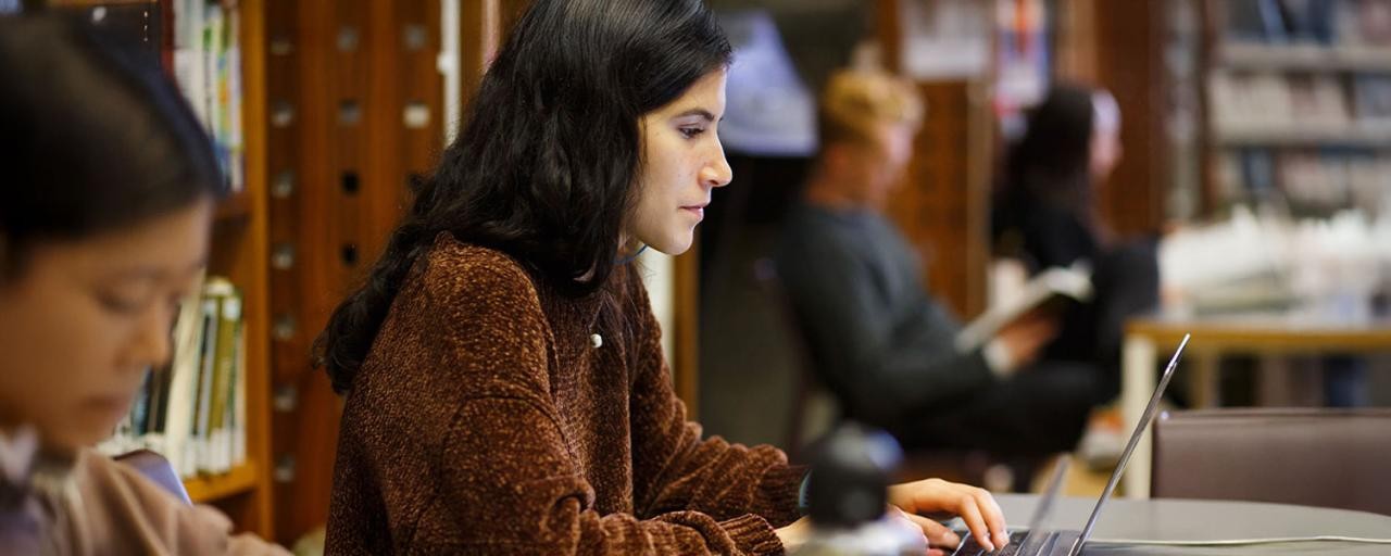 A woman in a cozy sweater focuses intently on her laptop in a library setting.