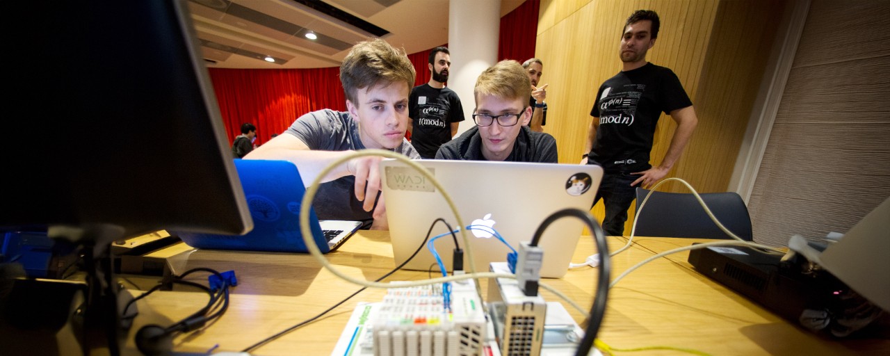 Two young men work closely together on a laptop surrounded by electronic components and cables at a table, while three others stand nearby observing in a brightly lit room with red curtains and wooden paneling.