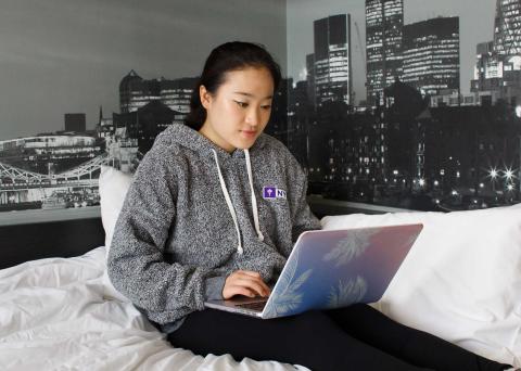 A person in a gray hoodie with a small NYU logo sits on a bed with white sheets, using a laptop.