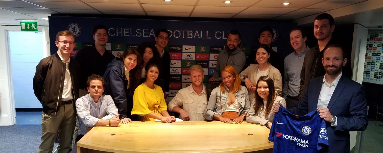 Fifteen NYU SPS students posing for a group photo in front of the Chelsea Football Club wall, smiling and representing the program during a site visit.