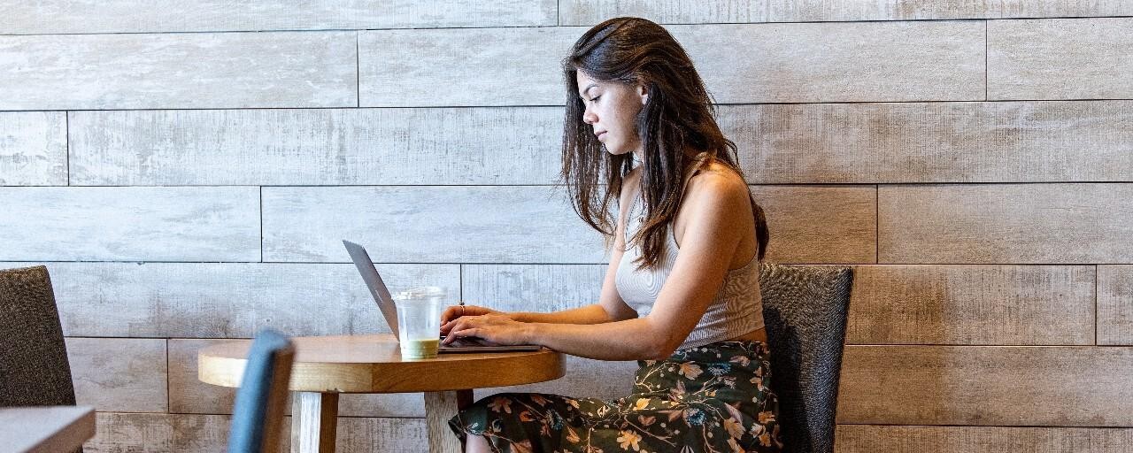 A woman sits in a cozy café, focused on her laptop. She wears a floral skirt and tank top. A drink and leather bag are on the table.
