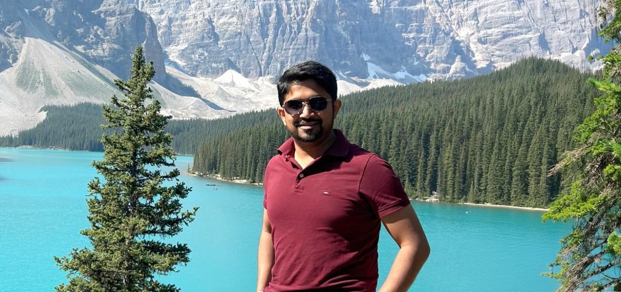 NYU SPS Division of Programs in Business alumnus Samarth Bedakihale poses for a photo in front of a scenic lake surrounded by trees and mountains.