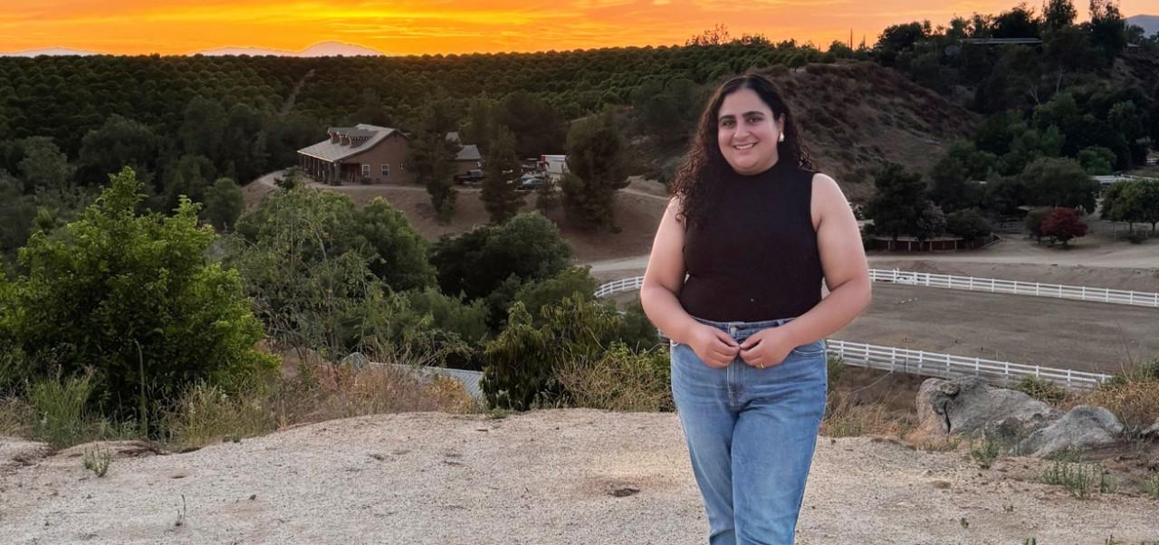 Sandra Khalil, Center for Global Affairs at NYU SPS alumnus, smiles for the camera while standing on a hill in front of a dramatic sunset.