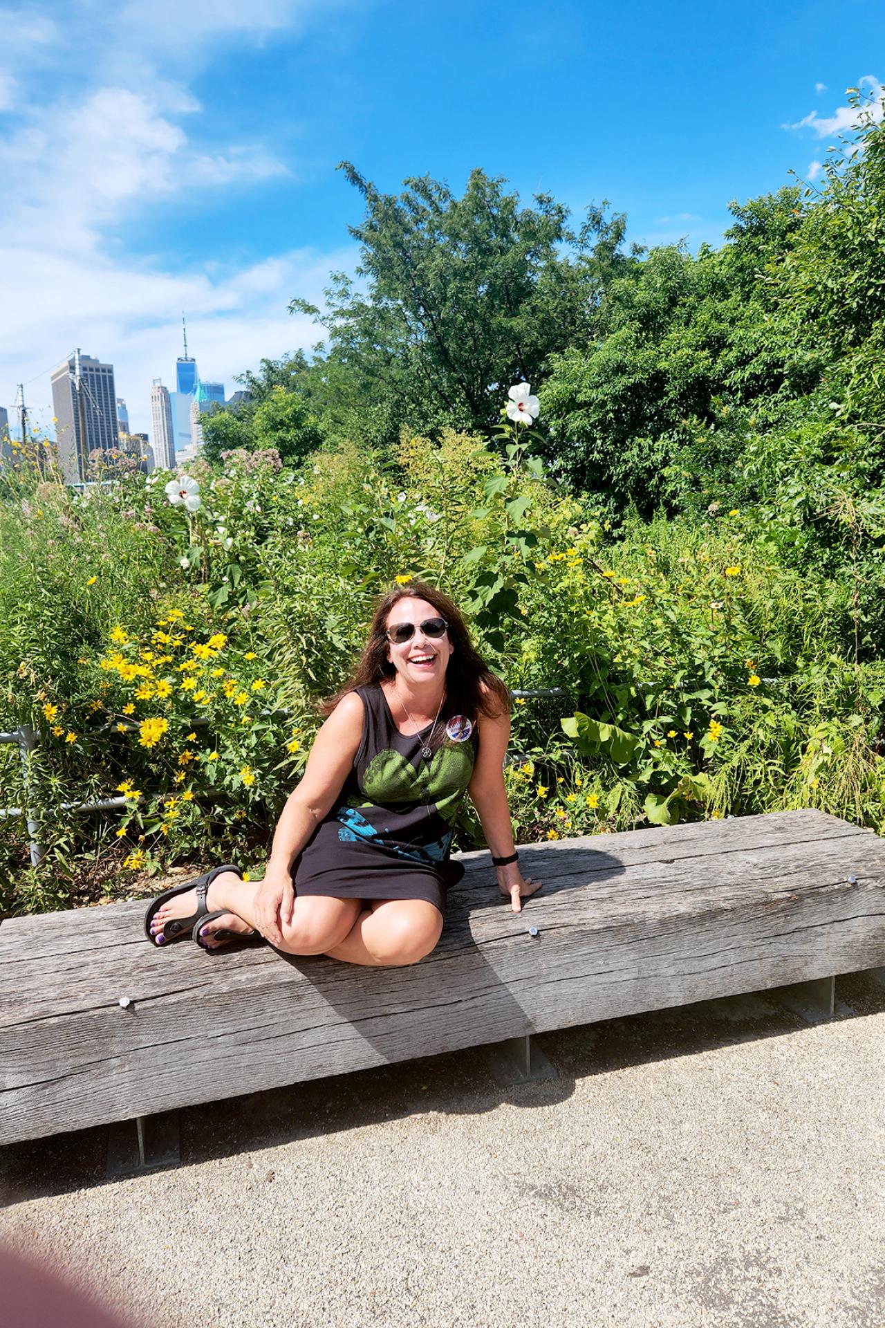 NYU SPS CE instructor Cathy Garrard smiles while sitting with her legs up on a park bench in an urban green space
