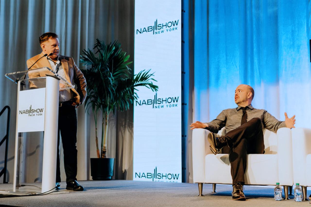 stands at a podium while another sits on a white chair, gesturing, both at a NAB Show in New York.