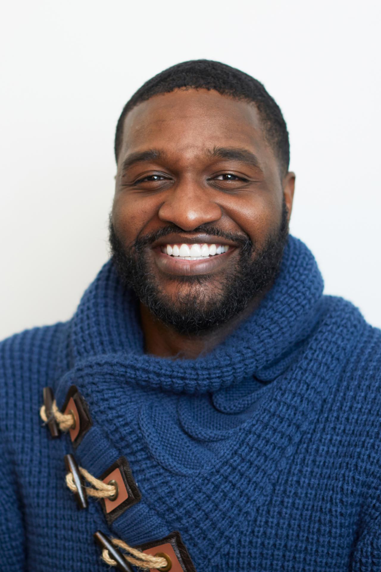 Eli Joseph smiles for a headshot while wearing a blue sweater in front of a white background