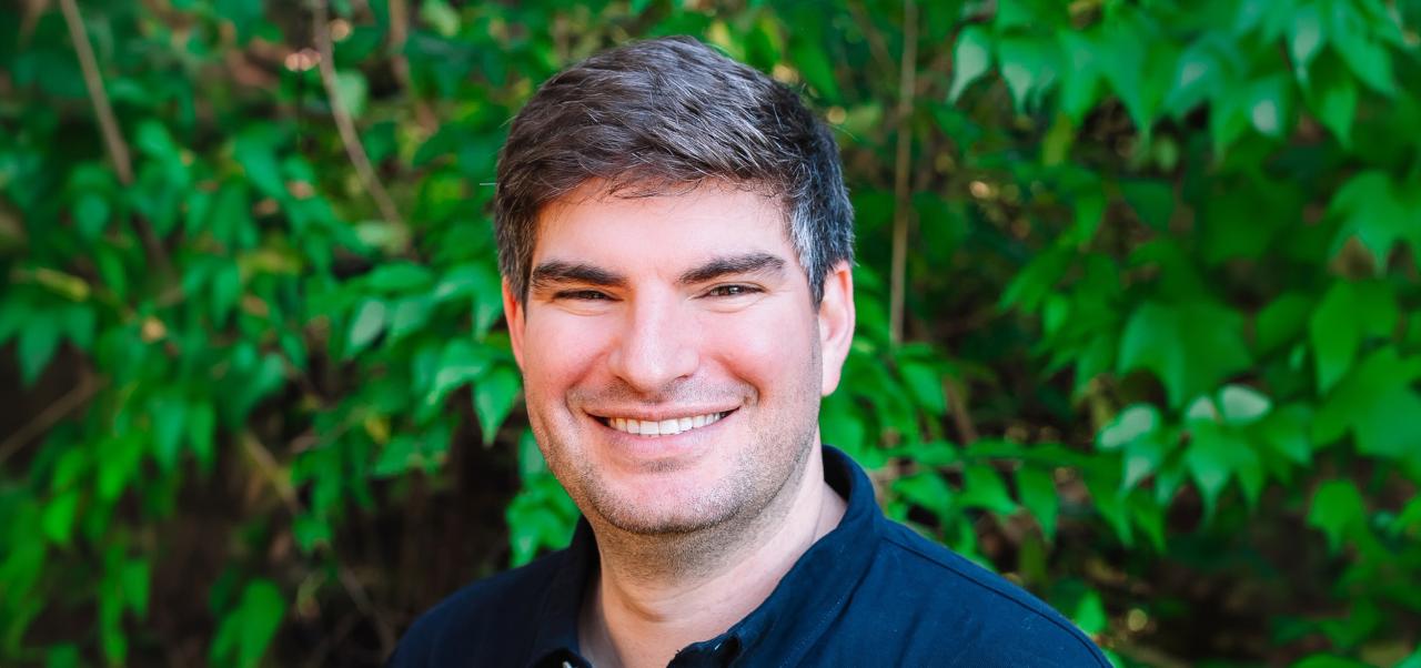 Jason Shames slightly smiles for a headshot wearing a navy blue blazer and a white shirt with a white backdrop.