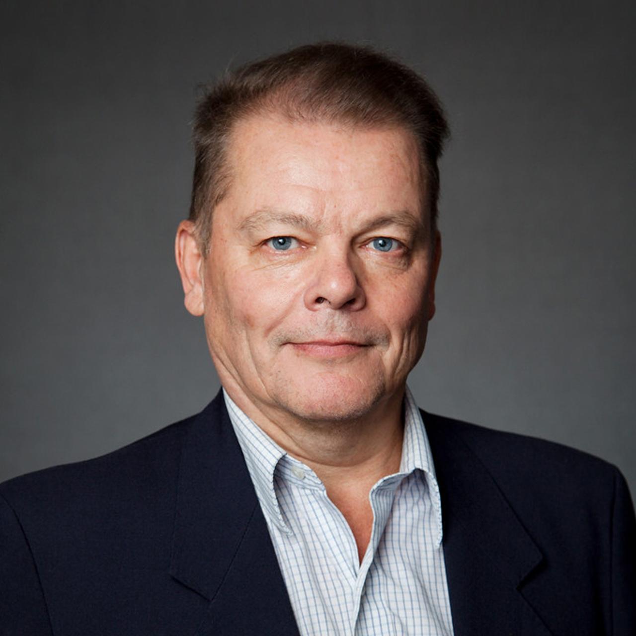 Jukka Laitamaki poses for a headshot in front of a grey background while wearing a black suit jacket and a blue grid-patterned shirt