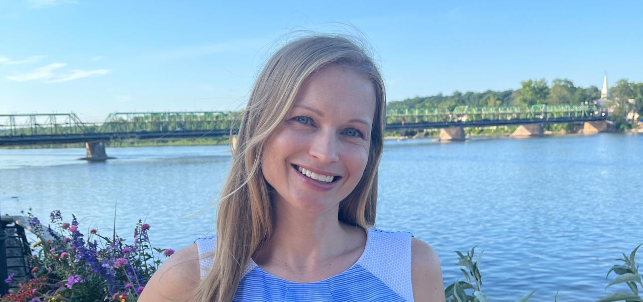 Mary Beth Altier smiles in a blue dress with a river and a bridge behind her.