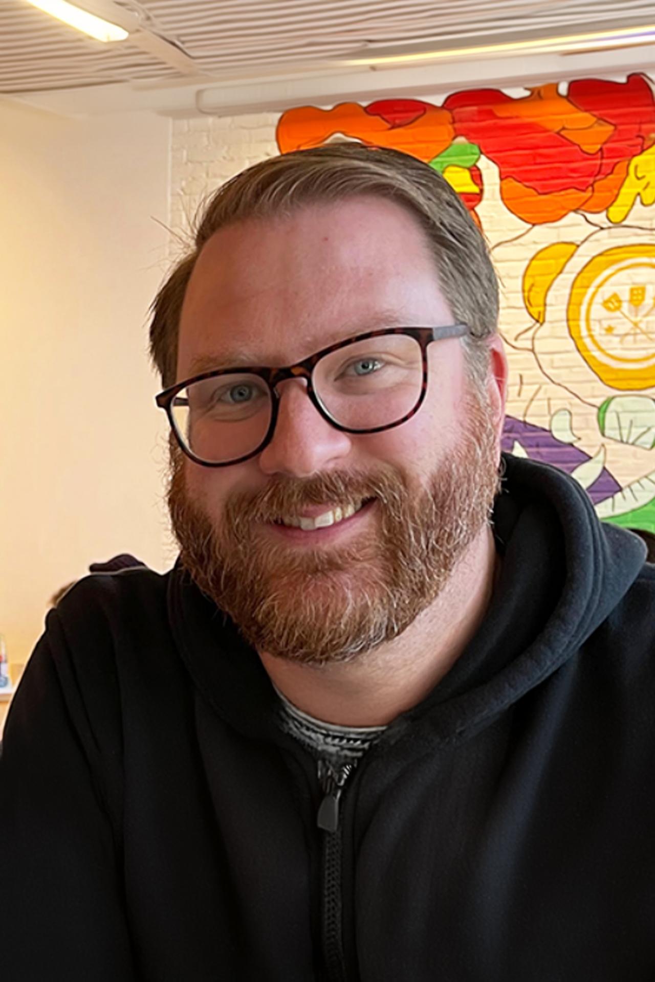 Patrick Gray smiles for a photo while wearing a black zippered jacket in front of a mural in the background