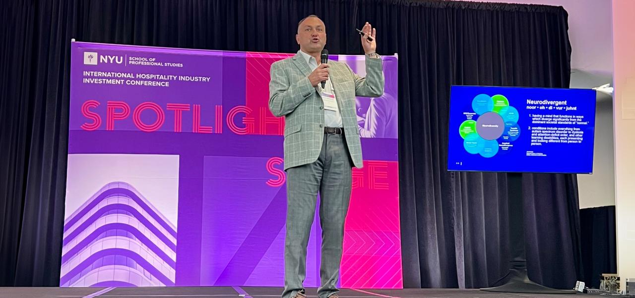 Recep Karaburun speaks with a microphone and one hand raised on a stage with a purple and pink NYU International Hospitality Industry Investment Conference Spotlight Stage banner behind him.