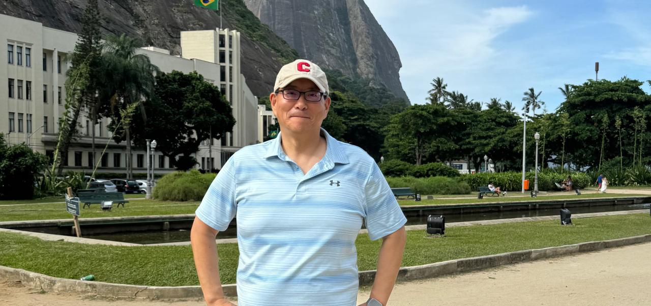In a tropical, urban setting, Steven Pae wears a blue polo shirt and a baseball cap and stands with his hands on his hips in front of an off-white building