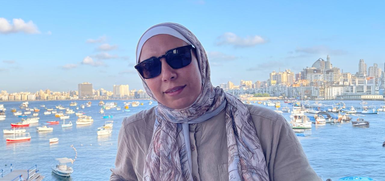  Yasmin Alkashef, Adjunct Instructor, Center for Publishing, Writing, and Media at NYU SPS, poses for a photo in front of a busy, gleaming, urban harbor full of watercraft.
