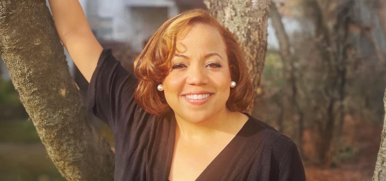 Cherese Blue wears a black shirt while smiling and leaning on a tree