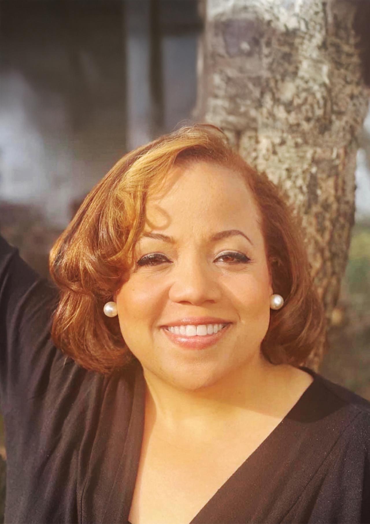 Cherese Blue wears a black shirt while smiling and leaning on a tree