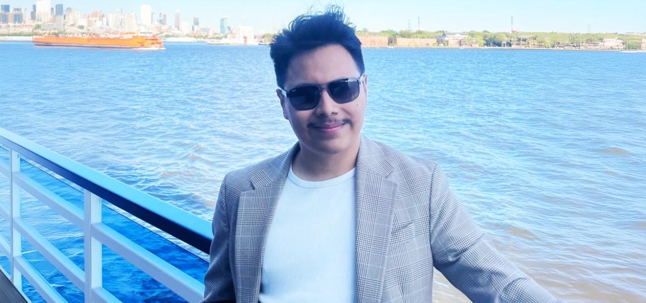Jonathan Hernandez Jimenez smiles for a headshot while on a boat with a river and a city scape in the background