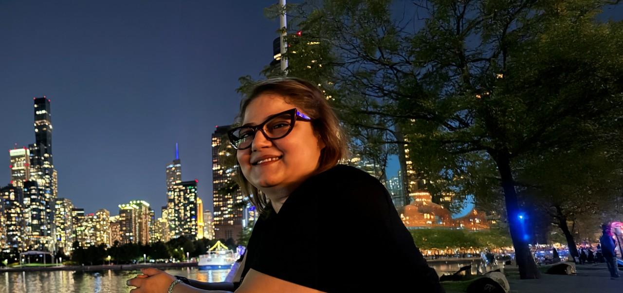 Reem Okar, student at the Center for Publishing and Applied Liberal Arts, NYU SPS smiles for the camera while dining at an outdoor restaurant with a view of the Manhattan skyline in the background.