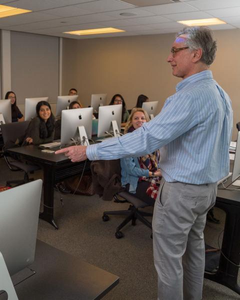 Man smiling and pointing to the left, rows of students in front of monitors looking towards the man