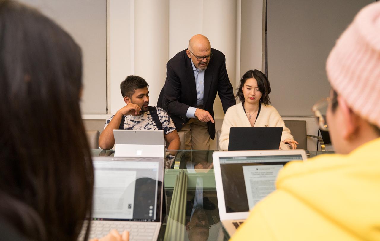 Man in back of room pointing and talking to one of four students on laptops