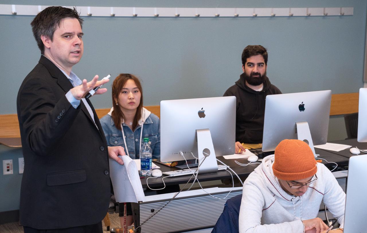 A classroom scene with a man in a black suit lecturing while students sit at tables with computers.