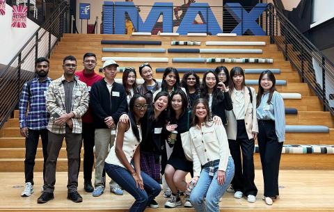 A group of students pose and smile on a staircase in front of a large IMAX sign.