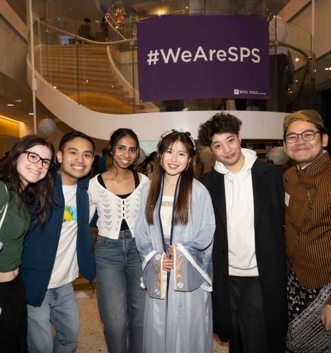 Six NYU SPS students smiling and posing in front of a '#WeAreSPS' sign displayed on the ledge of a staircase