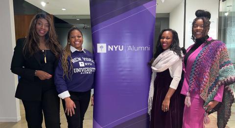 Four women smiling stand beside a purple "NYU Alumni" banner. They're dressed in business attire.