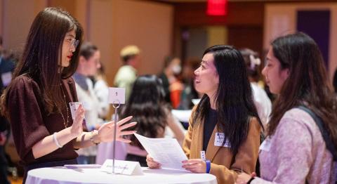 Three women engage in conversation at a networking event, around a tall table labeled "1." 
