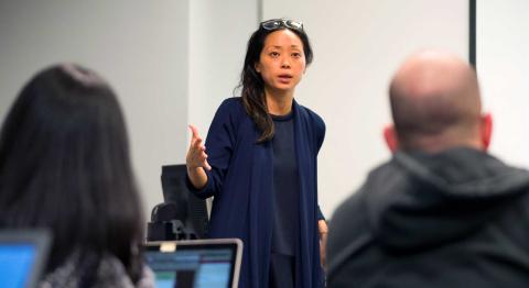 A woman in a blue cardigan speaks passionately at the front of a classroom. Two students with laptops listen.