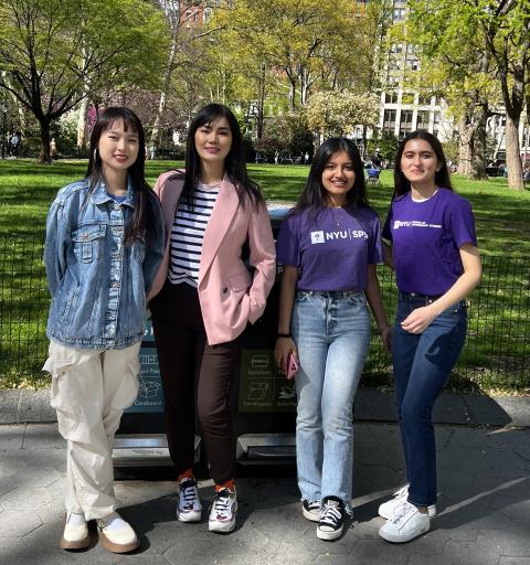 Four female scholarship students, two wearing purple NYU SPS shirts, pose for a photo in a park