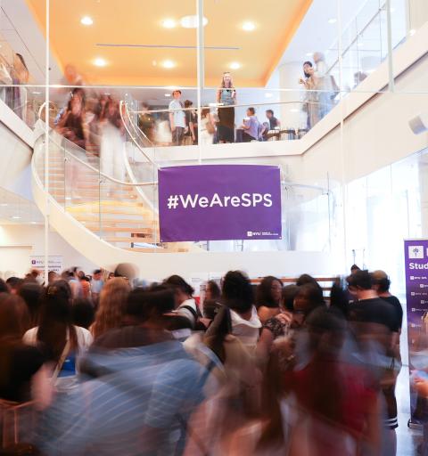 Crowded university lobby with students moving around, a purple "#WeAreSPS" banner hung above. Energetic atmosphere with blurred motion.