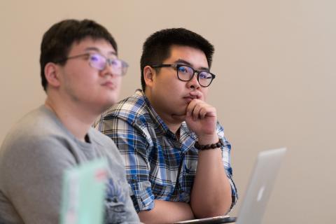 Two people seated indoors; one wearing glasses looks thoughtfully at a laptop during a meeting or class.