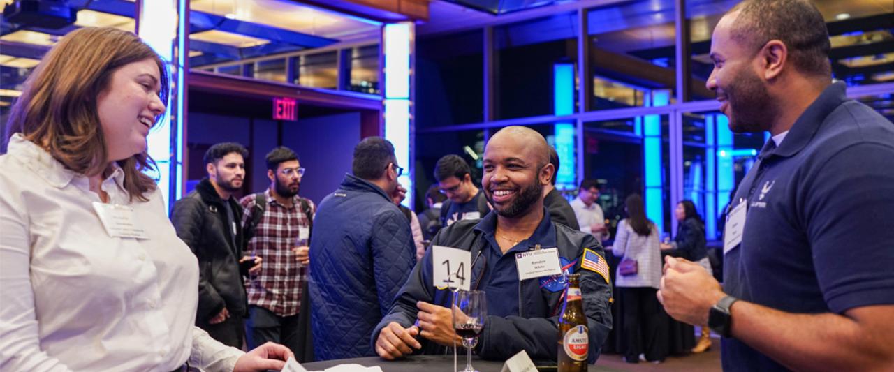 Three NYU SPS students gather at a table in the foreground and several others gather at tables in the background during a networking event.