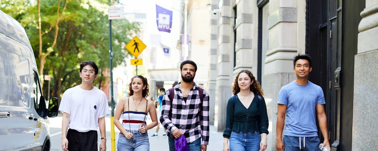 A group of five young adults walks confidently down a sunny city street. Behind them, NYU flags are visible, evoking a sense of academic life.